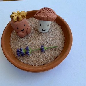 May include: A terracotta bowl filled with sand, featuring two small, smiling clay figures: a mushroom and a root vegetable with a golden crown. A sprig of lavender rests in the sand. The bowl is set against a white background.