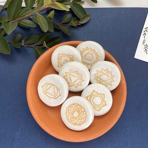 May include: Seven white round stones with gold chakra symbols on a terracotta plate. The stones are arranged in a circle with the symbols facing up.