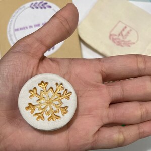 May include: A round, white, decorative item with a gold snowflake design. The item is held in a hand, with a small bag and a cardboard box visible in the background. The box has text that reads "Heavenly in the Airships".