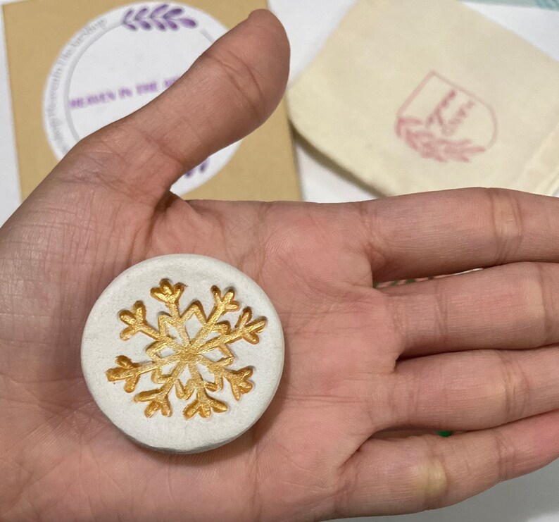 May include: A round, white, decorative item with a gold snowflake design. The item is held in a hand, with a small bag and a cardboard box visible in the background. The box has text that reads "Heavenly in the Airships".