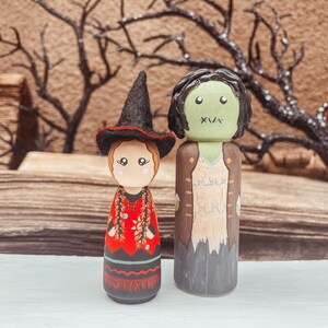 May include: A pair of hand-painted wooden peg dolls dressed as a witch and a zombie. The witch doll wears a black hat and a red and black dress. The zombie doll wears a brown jacket and has green skin.