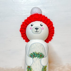 May include: A white wooden doll with a painted face and a red knitted hat with a white pom-pom. The doll is holding a green and red holly sprig.