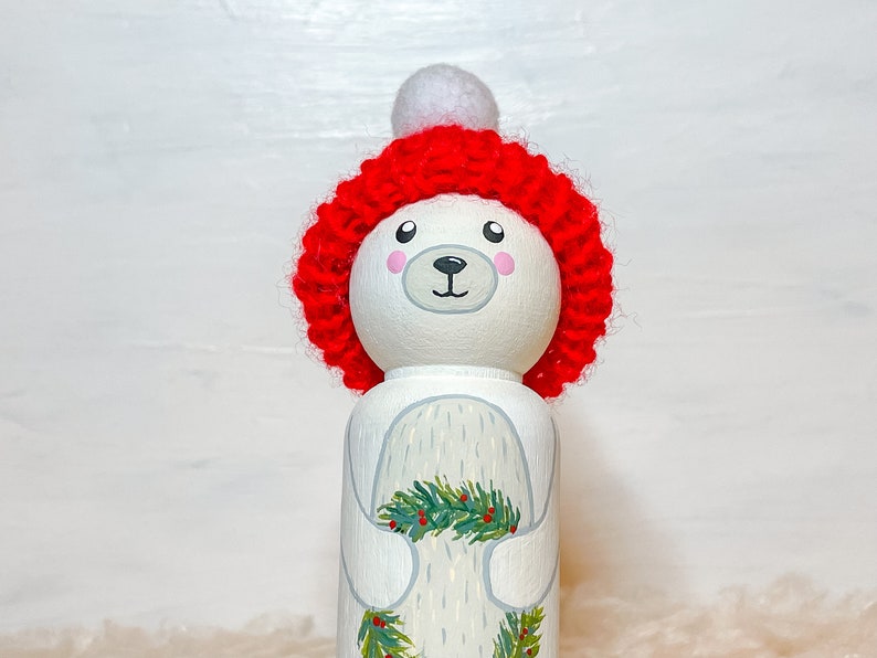 May include: A white wooden doll with a painted face and a red knitted hat with a white pom-pom. The doll is holding a green and red holly sprig.