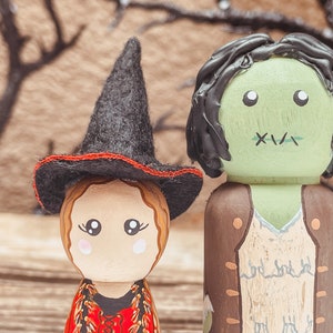 May include: Two wooden peg dolls dressed as Halloween characters. The doll on the left is a witch wearing a black hat with a red band and a red dress. The doll on the right is a green monster wearing a brown vest and a white shirt. The dolls are standing in front of a brown background.
