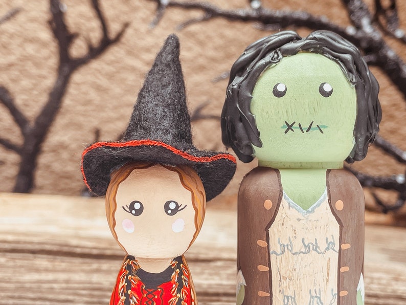 May include: Two wooden peg dolls dressed as Halloween characters. The doll on the left is a witch wearing a black hat with a red band and a red dress. The doll on the right is a green monster wearing a brown vest and a white shirt. The dolls are standing in front of a brown background.