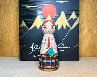 May include: A hand-painted wooden doll with red hair, wearing glasses, a cream sweater with red flowers, a plaid skirt, and holding a book titled "The Lord of the Rings".
