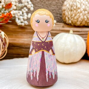 Op de afbeelding: Een houten pegdol met blond haar, gekleed in een paarse en gouden jurk met een hemels patroon. De pop staat op een houten oppervlak met pompoenen op de achtergrond.