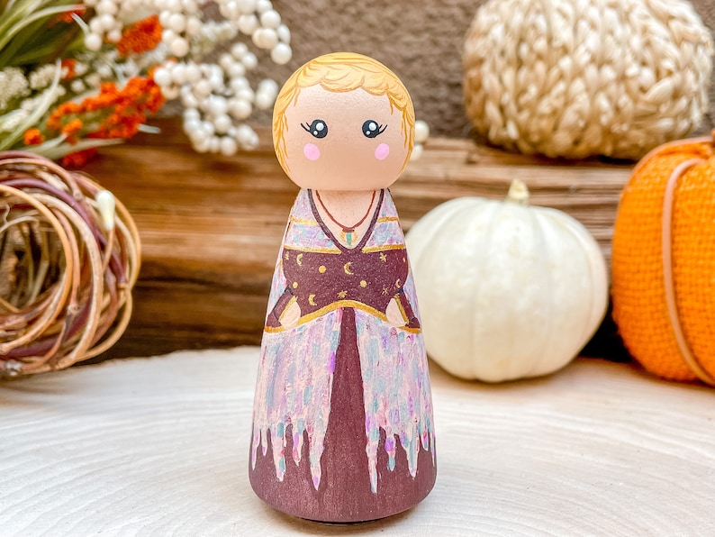 Op de afbeelding: Een houten pegdol met blond haar, gekleed in een paarse en gouden jurk met een hemels patroon. De pop staat op een houten oppervlak met pompoenen op de achtergrond.