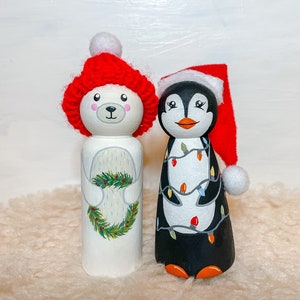 May include: A white polar bear and a black penguin wearing red Santa hats. The polar bear is holding a green wreath and the penguin is decorated with colorful Christmas lights.
