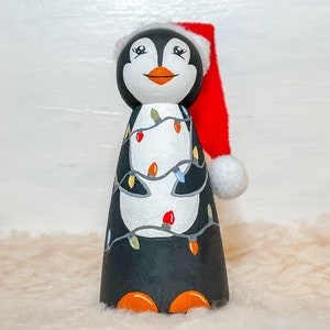 May include: A hand-painted wooden penguin figurine wearing a red Santa hat with a white pom-pom. The penguin is decorated with colorful Christmas lights.