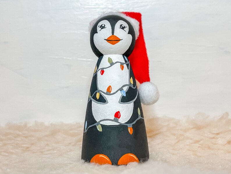 May include: A hand-painted wooden penguin figurine wearing a red Santa hat with a white pom-pom. The penguin is decorated with colorful Christmas lights.