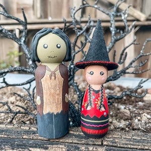 May include: Two hand-painted wooden peg dolls dressed in Halloween costumes. The doll on the left is a zombie with green skin and a brown and black outfit. The doll on the right is a witch with a black hat and a red and black dress.