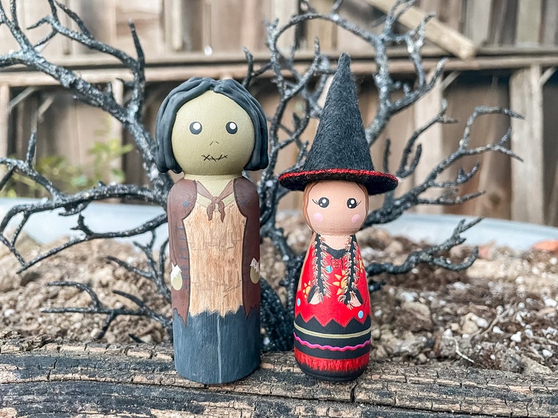 May include: Two hand-painted wooden peg dolls dressed in Halloween costumes. The doll on the left is a zombie with green skin and a brown and black outfit. The doll on the right is a witch with a black hat and a red and black dress.