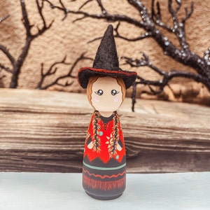 May include: A wooden doll dressed as a witch with a black pointed hat, red and black dress, and a long braid of hair. The doll is standing on a wooden surface.