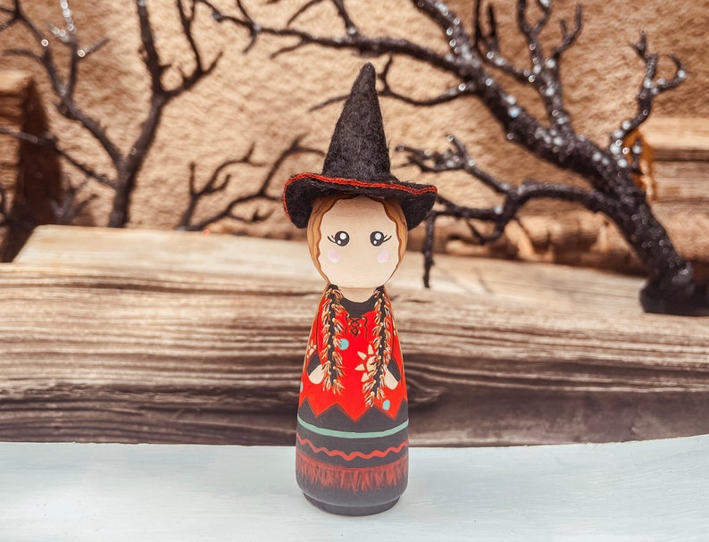 May include: A wooden doll dressed as a witch with a black pointed hat, red and black dress, and a long braid of hair. The doll is standing on a wooden surface.