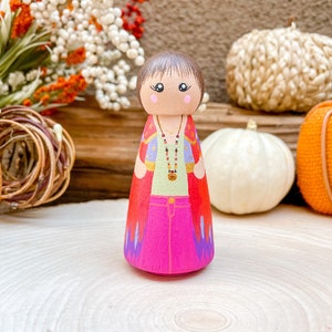 Op de afbeelding: Een houten pegdoll met bruin haar, geschilderd met een groen shirt, een roze broek en een rode en paarse cape. De pop draagt een gouden ketting met rode kralen en een gouden hanger.
