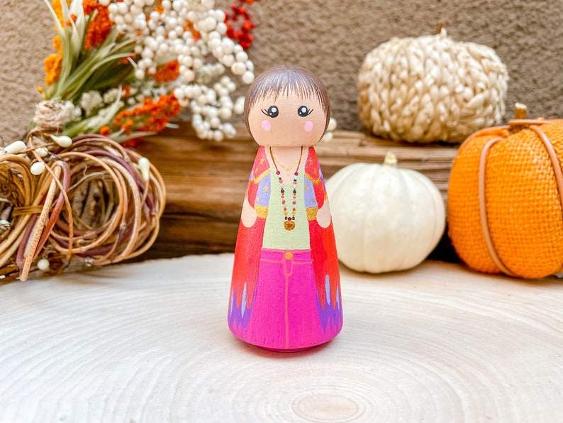 Op de afbeelding: Een houten pegdoll met bruin haar, geschilderd met een groen shirt, een roze broek en een rode en paarse cape. De pop draagt een gouden ketting met rode kralen en een gouden hanger.