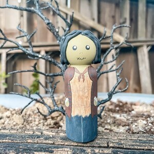May include: A hand-painted wooden doll with a stitched mouth, brown hair, and a brown and tan outfit. The doll is standing on a wooden surface.