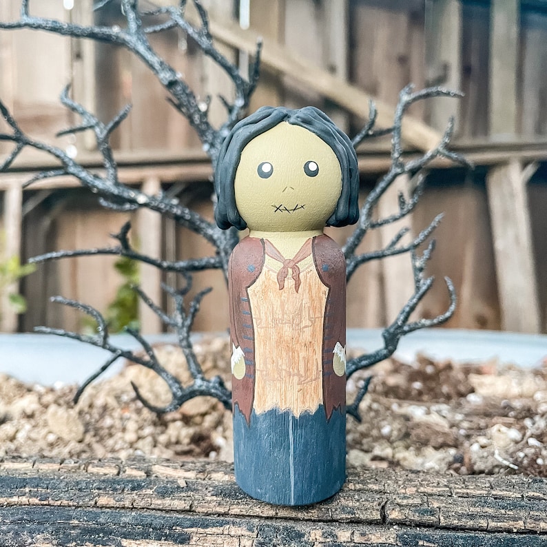May include: A hand-painted wooden doll with a stitched mouth, brown hair, and a brown and tan outfit. The doll is standing on a wooden surface.