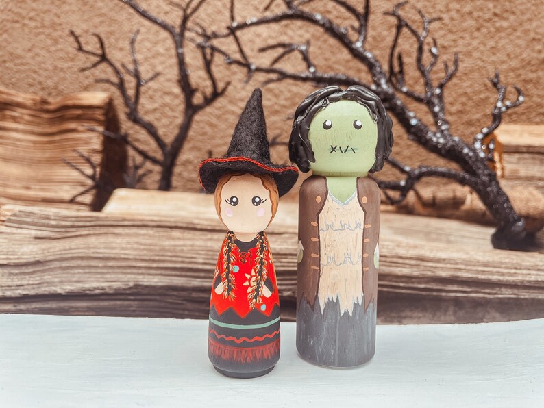 May include: A pair of hand-painted wooden peg dolls dressed as a witch and a zombie. The witch doll wears a black hat and a red and black dress. The zombie doll wears a brown jacket and has green skin.