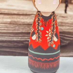 May include: A hand-painted wooden doll with a red dress, black and white geometric patterns, and gold and brown fringe. The doll has a painted face with brown hair and blue eyes.