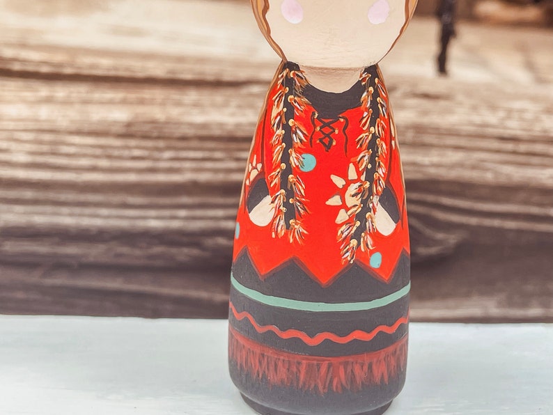 May include: A hand-painted wooden doll with a red dress, black and white geometric patterns, and gold and brown fringe. The doll has a painted face with brown hair and blue eyes.