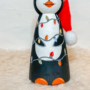May include: A hand-painted wooden penguin figurine wearing a red Santa hat with a white pom-pom. The penguin is decorated with a string of colorful Christmas lights.
