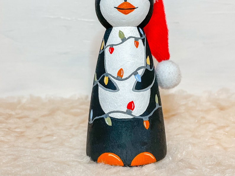 May include: A hand-painted wooden penguin figurine wearing a red Santa hat with a white pom-pom. The penguin is decorated with a string of colorful Christmas lights.