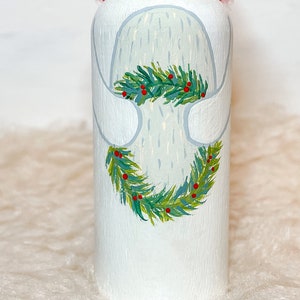 May include: White painted wooden ornament with a green and red wreath painted around it. The wreath has green leaves and red berries.