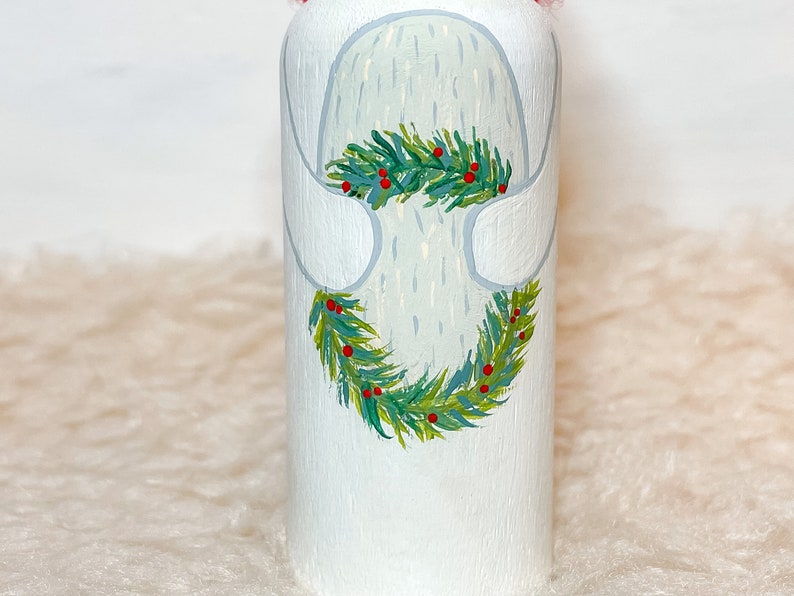 May include: White painted wooden ornament with a green and red wreath painted around it. The wreath has green leaves and red berries.