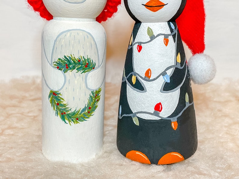 May include: A white polar bear peg doll with a green wreath and red berries, and a black and white penguin peg doll wearing a red Santa hat with white trim and white lights.