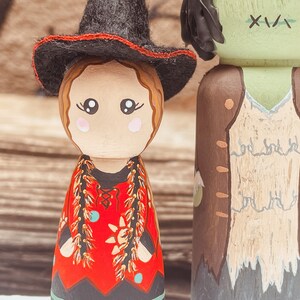 May include: A pair of wooden peg dolls dressed as a witch and a zombie. The witch doll is wearing a black hat with a red band, a red dress with gold trim, and a black skirt. The zombie doll is wearing a brown vest over a white shirt, and has green skin with stitches on its face.
