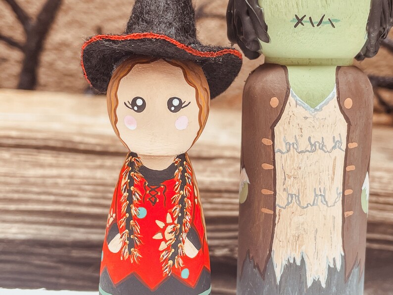 May include: A pair of wooden peg dolls dressed as a witch and a zombie. The witch doll is wearing a black hat with a red band, a red dress with gold trim, and a black skirt. The zombie doll is wearing a brown vest over a white shirt, and has green skin with stitches on its face.