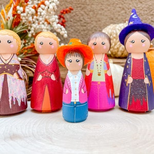 Op de afbeelding: Vijf houten pegdolle, elk met een andere outfit en haarstijl. De poppen zijn geschilderd in verschillende kleuren, waaronder rood, blauw, roze en paars. De poppen staan op een houten oppervlak.