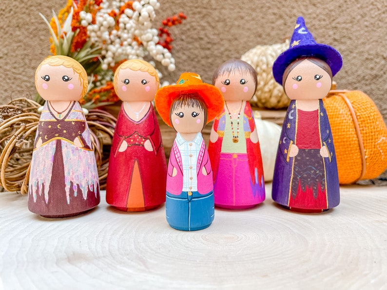 Op de afbeelding: Vijf houten pegdolle, elk met een andere outfit en haarstijl. De poppen zijn geschilderd in verschillende kleuren, waaronder rood, blauw, roze en paars. De poppen staan op een houten oppervlak.