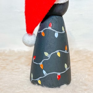 May include: A black wooden Christmas tree ornament with a red Santa hat and white trim. The tree is decorated with a string of colorful lights.