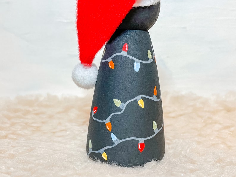 May include: A black wooden Christmas tree ornament with a red Santa hat and white trim. The tree is decorated with a string of colorful lights.