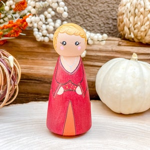 Op de afbeelding: Een houten pegdoll met blond haar, gekleed in een rode jurk met gouden accenten en een zilveren ketting. De pop staat op een houten oppervlak.