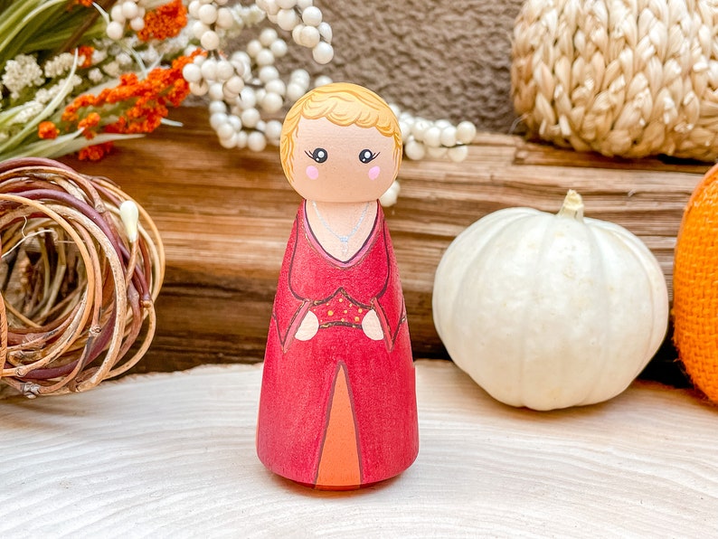 Op de afbeelding: Een houten pegdoll met blond haar, gekleed in een rode jurk met gouden accenten en een zilveren ketting. De pop staat op een houten oppervlak.