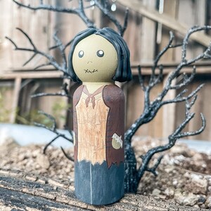 May include: A hand-painted wooden doll with a green face, black hair, and a brown and tan outfit. The doll is wearing a brown jacket with a tan shirt underneath. The doll has a stitched mouth and eyes.