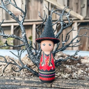 May include: A wooden doll with a black witch hat and a red, black, and yellow patterned dress. The doll is standing on a wooden surface.