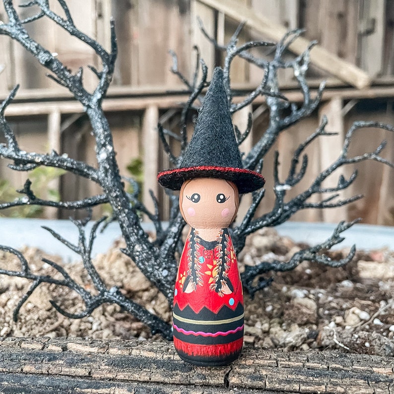 May include: A wooden doll with a black witch hat and a red, black, and yellow patterned dress. The doll is standing on a wooden surface.