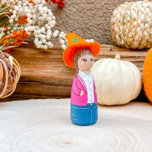 Op de afbeelding: Een handgeschilderde houten pop met een roze jas, blauwe jeans en een oranje heksenhoed. De pop staat op een houten oppervlak met pompoenen en herfstdecoratie op de achtergrond.