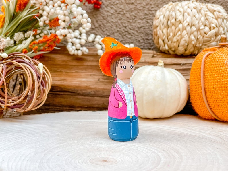 Op de afbeelding: Een handgeschilderde houten pop met een roze jas, blauwe jeans en een oranje heksenhoed. De pop staat op een houten oppervlak met pompoenen en herfstdecoratie op de achtergrond.