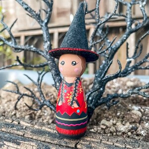 May include: A wooden doll with a black witch hat and a red, black, and gold patterned dress. The doll is standing on a wooden surface.