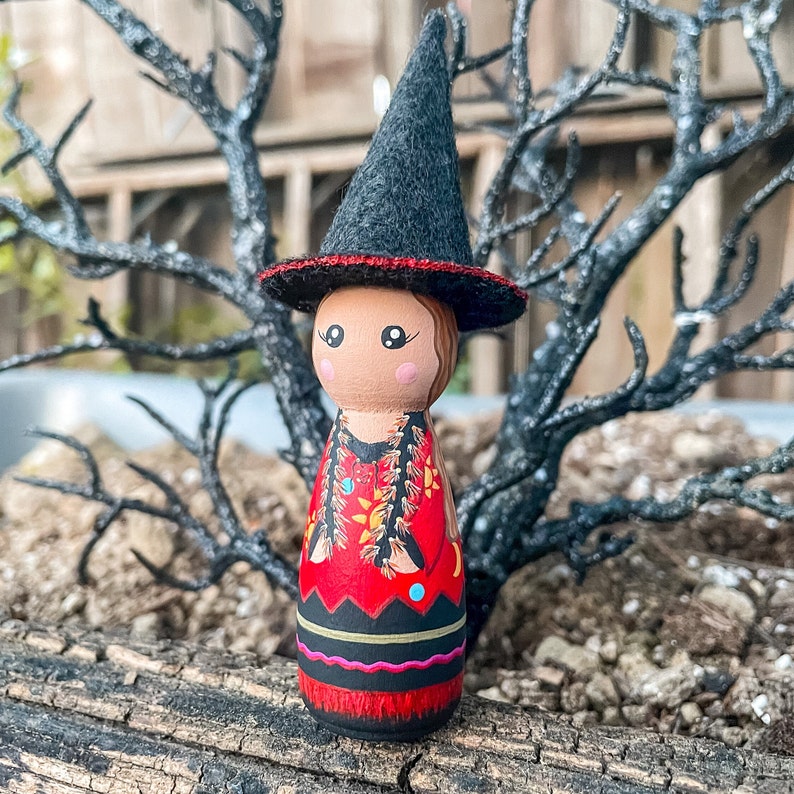 May include: A wooden doll with a black witch hat and a red, black, and gold patterned dress. The doll is standing on a wooden surface.