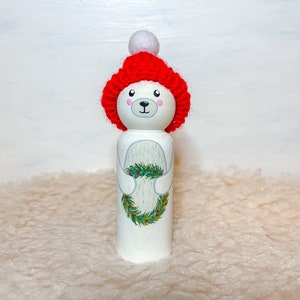 May include: A white wooden doll painted to look like a polar bear wearing a red knit hat with a white pom-pom. The doll is holding a green wreath with red berries.