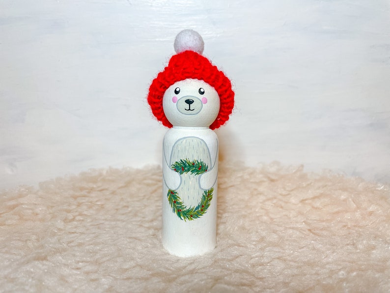 May include: A white wooden doll painted to look like a polar bear wearing a red knit hat with a white pom-pom. The doll is holding a green wreath with red berries.