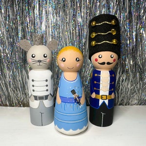 Pode incluir: Três bonecas de madeira pintadas à mão, um rato, uma menina com um vestido azul e um soldado com um uniforme azul e dourado. As bonecas estão todas em pé sobre uma superfície branca.