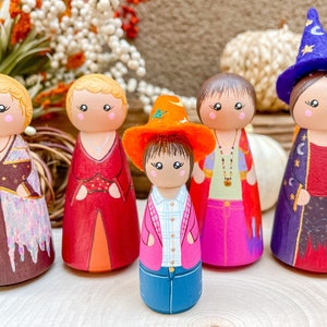 Op de afbeelding: Vijf houten pegdolle met beschilderde gezichten en kleding. De poppen zijn gekleed in verschillende outfits, waaronder een rode jurk, een paarse robe, een roze jurk, een blauw shirt en jeans, en een bruine jurk met een gouden ketting. De poppen staan op een houten oppervlak.